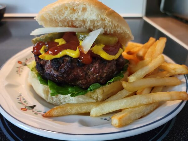 Grilled steak burger (hamburger) on a fresh bun with onions, lettuce, ketchup, pickles, and mustard, with fries on the side, on a dinner plate.