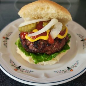 Grilled steak burger (hamburger) on a fresh bun with onions, lettuce, ketchup, pickles, and mustard, on a dinner plate.